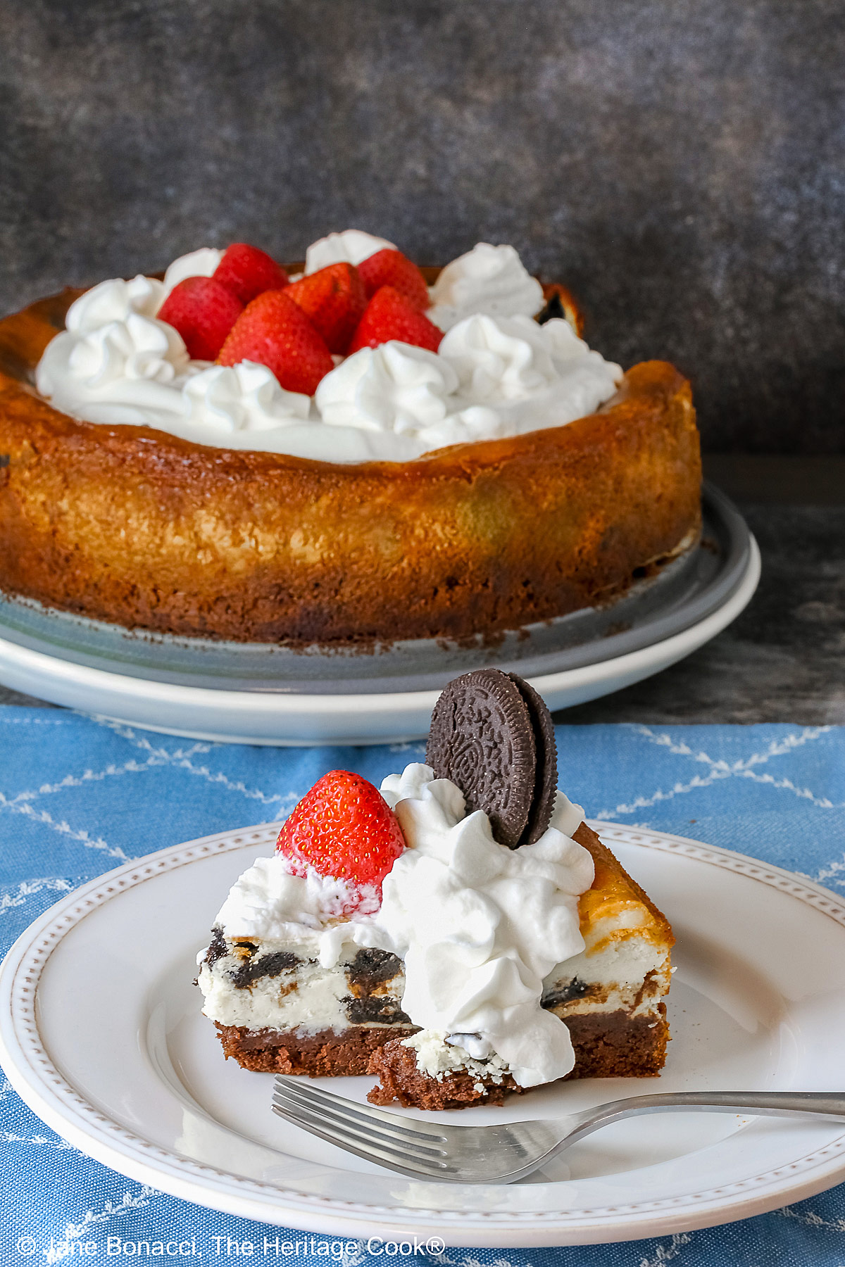 Whole cheesecake behind a slice on a white plate; A slice of Scrumptious Oreo Brownie Cheesecake, topped with freshly made whipped cream, a whole Oreo cookie, and a whole strawberry, with a sprig of mint alongside © 2026 Jane Bonacci, The Heritage Cook.