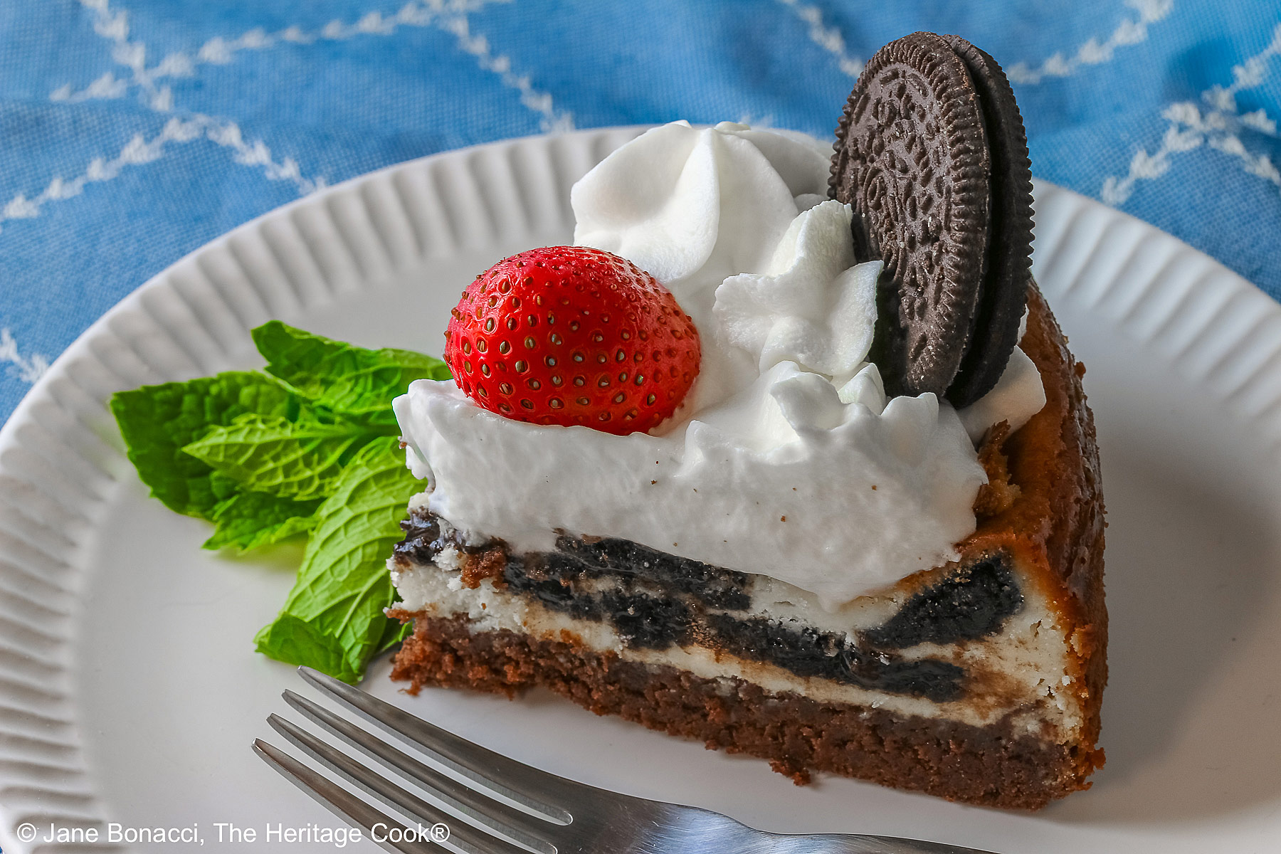 A slice of Scrumptious Oreo Brownie Cheesecake, topped with freshly made whipped cream, a whole Oreo cookie, and a whole strawberry, with a sprig of mint alongside © 2026 Jane Bonacci, The Heritage Cook.
