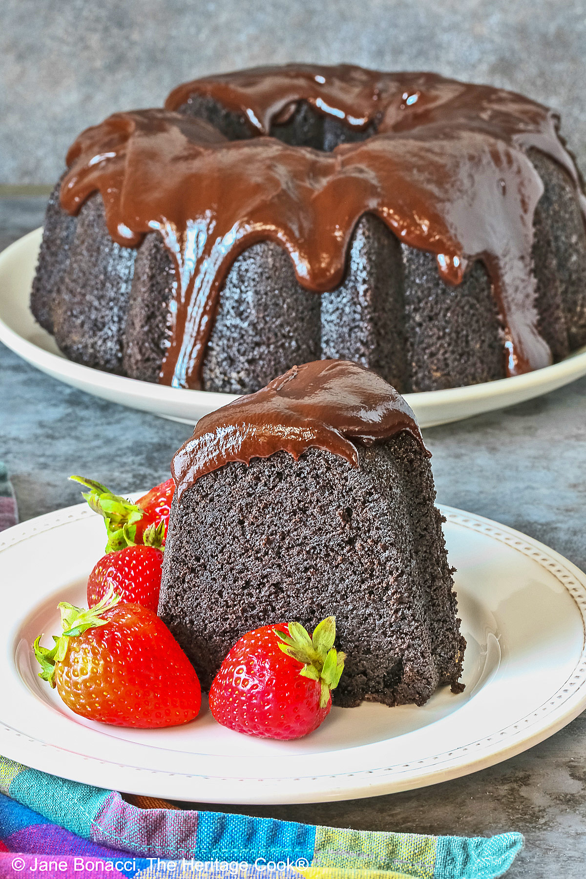 Super Simple Brownie Bundt Cake topped with glistening chocolate ganache glaze, most photos are a slice on a cream-colored plate, surrounded by whole strawberries with a fork on the opposite side, sitting on a grey background, a multi-colored cloth on the left and some with the whole cake behind © 2026 Jane Bonacci, The Heritage Cook.