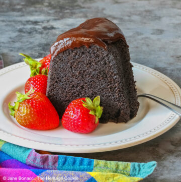 Super Simple Brownie Bundt Cake topped with glistening chocolate ganache glaze, most photos are a slice on a cream-colored plate, surrounded by whole strawberries with a fork on the opposite side, sitting on a grey background, a multi-colored cloth on the left and some with the whole cake behind © 2026 Jane Bonacci, The Heritage Cook.