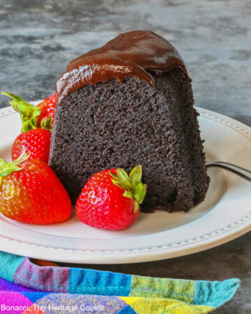 Super Simple Brownie Bundt Cake topped with glistening chocolate ganache glaze, most photos are a slice on a cream-colored plate, surrounded by whole strawberries with a fork on the opposite side, sitting on a grey background, a multi-colored cloth on the left and some with the whole cake behind © 2026 Jane Bonacci, The Heritage Cook.