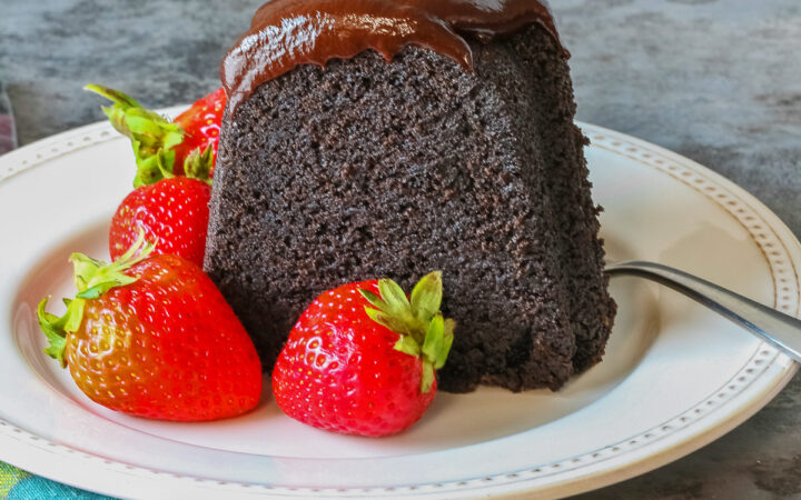 Super Simple Brownie Bundt Cake topped with glistening chocolate ganache glaze, most photos are a slice on a cream-colored plate, surrounded by whole strawberries with a fork on the opposite side, sitting on a grey background, a multi-colored cloth on the left and some with the whole cake behind © 2026 Jane Bonacci, The Heritage Cook.