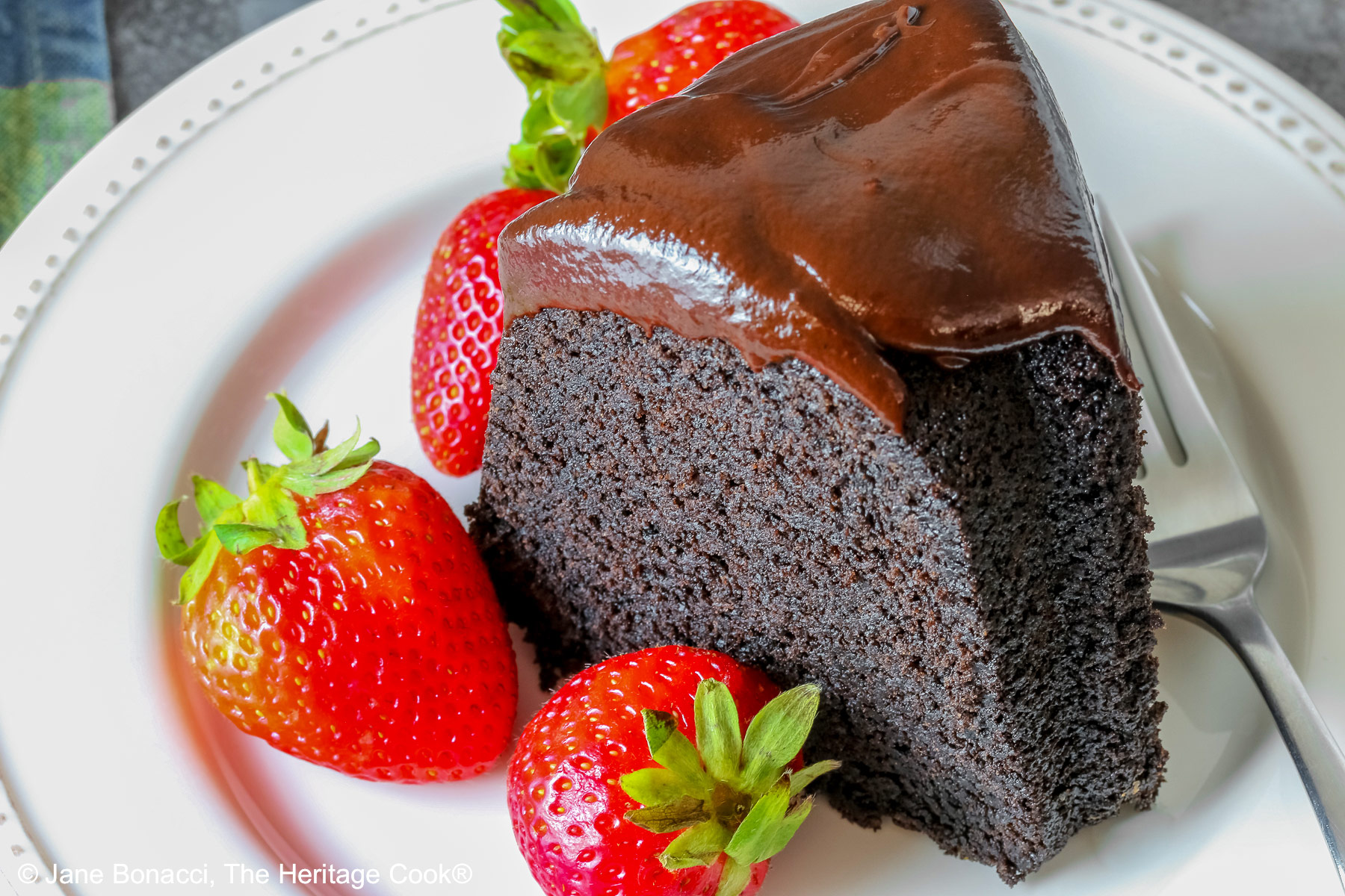 Super Simple Brownie Bundt Cake topped with glistening chocolate ganache glaze, most photos are a slice on a cream-colored plate, surrounded by whole strawberries with a fork on the opposite side, sitting on a grey background, a multi-colored cloth on the left and some with the whole cake behind © 2026 Jane Bonacci, The Heritage Cook.