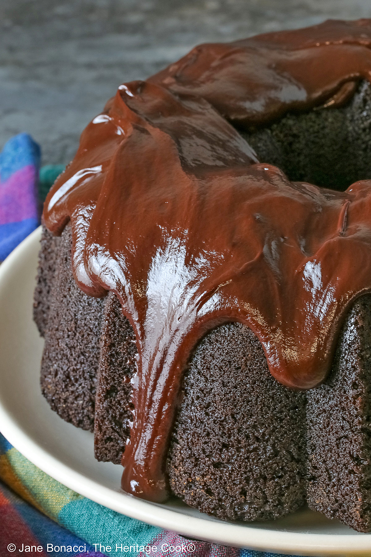 Super Simple Brownie Bundt Cake topped with glistening chocolate ganache glaze, most photos are a slice on a cream-colored plate, surrounded by whole strawberries with a fork on the opposite side, sitting on a grey background, a multi-colored cloth on the left and some with the whole cake behind © 2026 Jane Bonacci, The Heritage Cook.