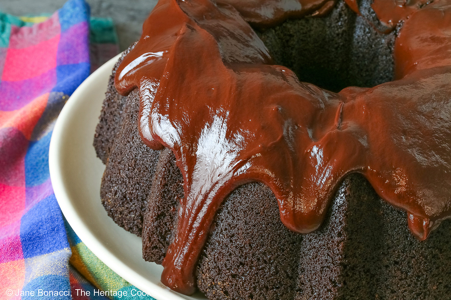 Super Simple Brownie Bundt Cake topped with glistening chocolate ganache glaze, most photos are a slice on a cream-colored plate, surrounded by whole strawberries with a fork on the opposite side, sitting on a grey background, a multi-colored cloth on the left and some with the whole cake behind © 2026 Jane Bonacci, The Heritage Cook.