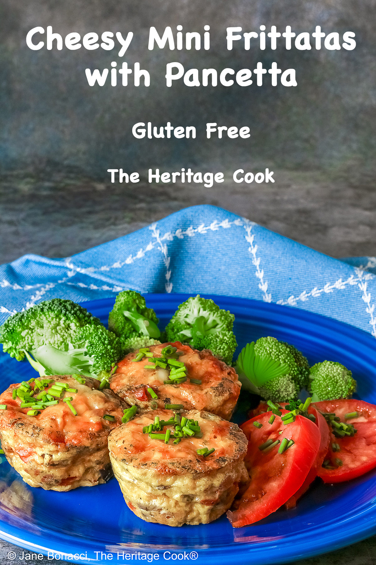 3 Cheesy Mini Frittatas with Pancetta on a blue plate with slices of red tomato and green broccoli beside them, with a blue and white cloth behind, sitting on a mottled gray background © 2026 Jane Bonacci, The Heritage Cook.