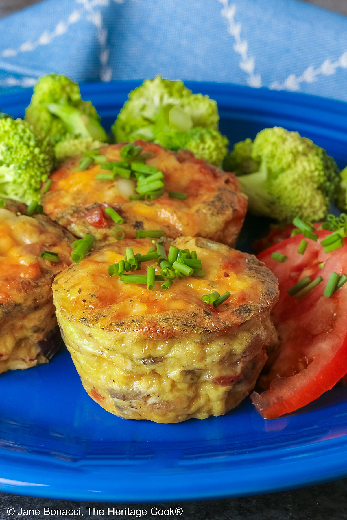 3 Cheesy Mini Frittatas with Pancetta on a blue plate with slices of red tomato and green broccoli beside them, with a blue and white cloth behind, sitting on a mottled gray background © 2026 Jane Bonacci, The Heritage Cook.