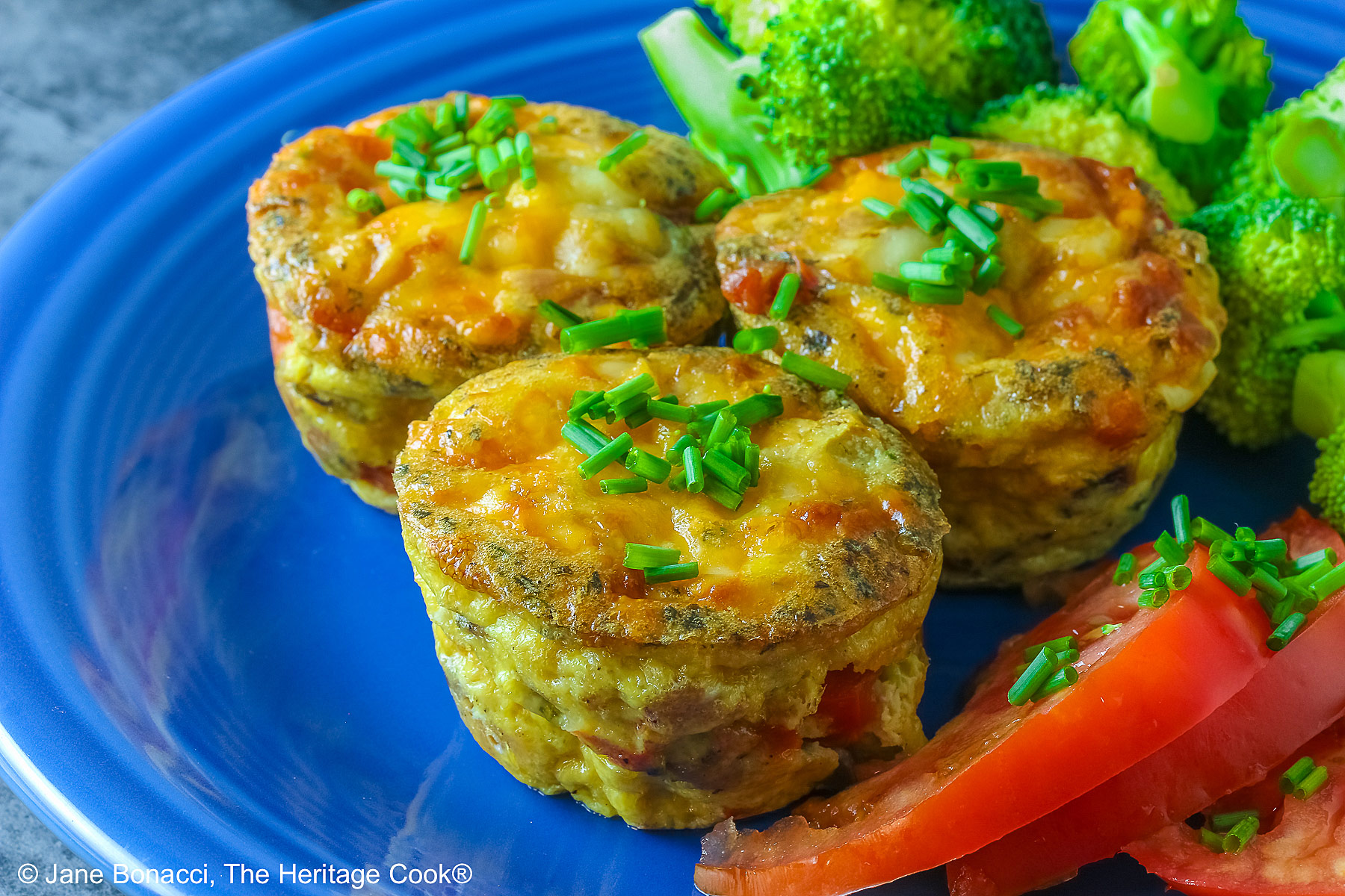 3 Cheesy Mini Frittatas with Pancetta on a blue plate with slices of red tomato and green broccoli beside them, with a blue and white cloth behind, sitting on a mottled gray background © 2026 Jane Bonacci, The Heritage Cook.