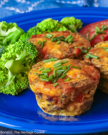 3 Cheesy Mini Frittatas with Pancetta on a blue plate with slices of red tomato and green broccoli beside them, with a blue and white cloth behind, sitting on a mottled gray background © 2026 Jane Bonacci, The Heritage Cook.