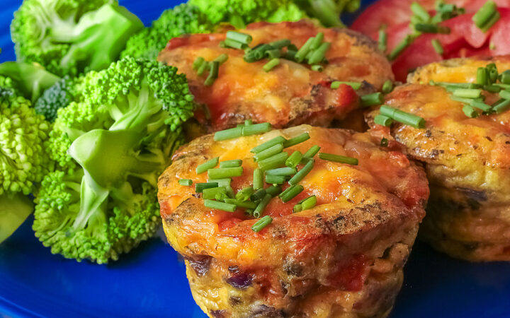 3 Cheesy Mini Frittatas with Pancetta on a blue plate with slices of red tomato and green broccoli beside them, with a blue and white cloth behind, sitting on a mottled gray background © 2026 Jane Bonacci, The Heritage Cook.