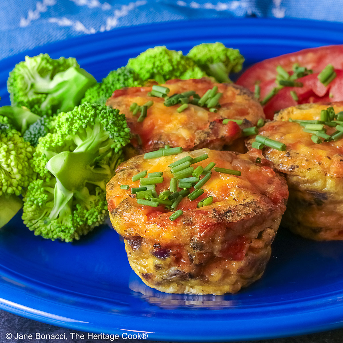 3 Cheesy Mini Frittatas with Pancetta on a blue plate with slices of red tomato and green broccoli beside them, with a blue and white cloth behind, sitting on a mottled gray background © 2026 Jane Bonacci, The Heritage Cook.