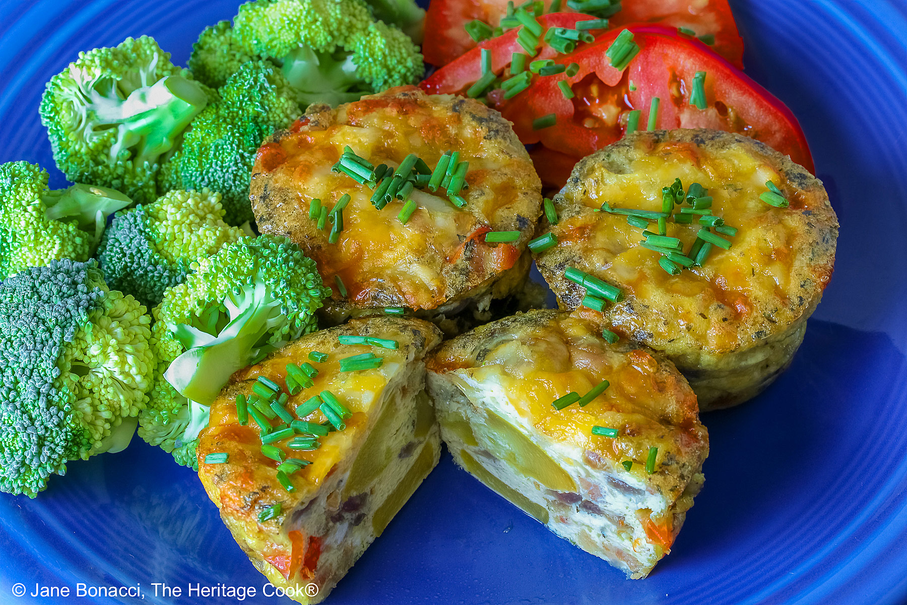 3 Cheesy Mini Frittatas with Pancetta on a blue plate with slices of red tomato and green broccoli beside them, with a blue and white cloth behind, sitting on a mottled gray background © 2026 Jane Bonacci, The Heritage Cook.
