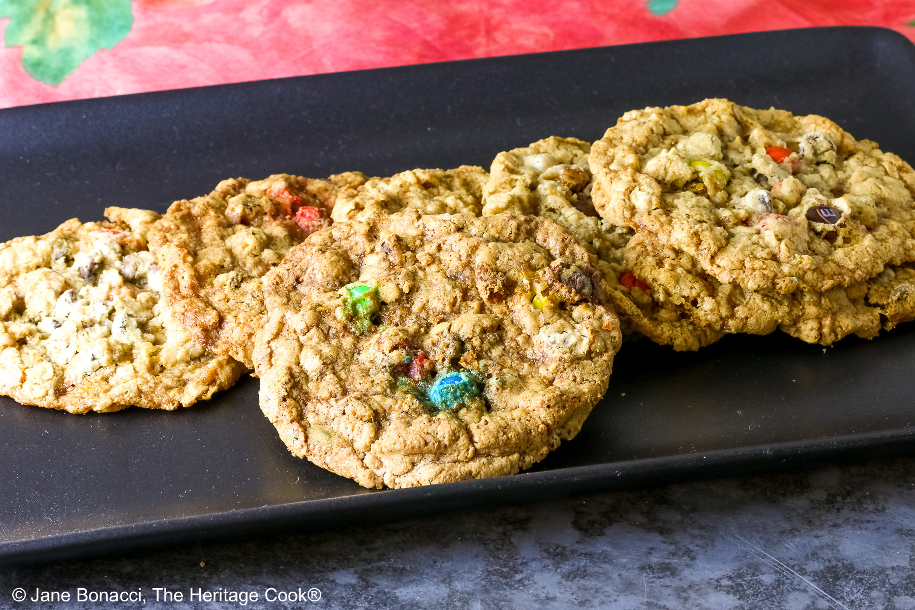 Large oatmeal cookies studded with all kinds of add-ins like coconut, pecans, chocolate chips, peanut butter chips, and M&M candies, laid out on a black rectangular platter with a deep pink cloth in the back and sitting on a mottled gray background © 2026 Jane Bonacci, The Heritage Cook.