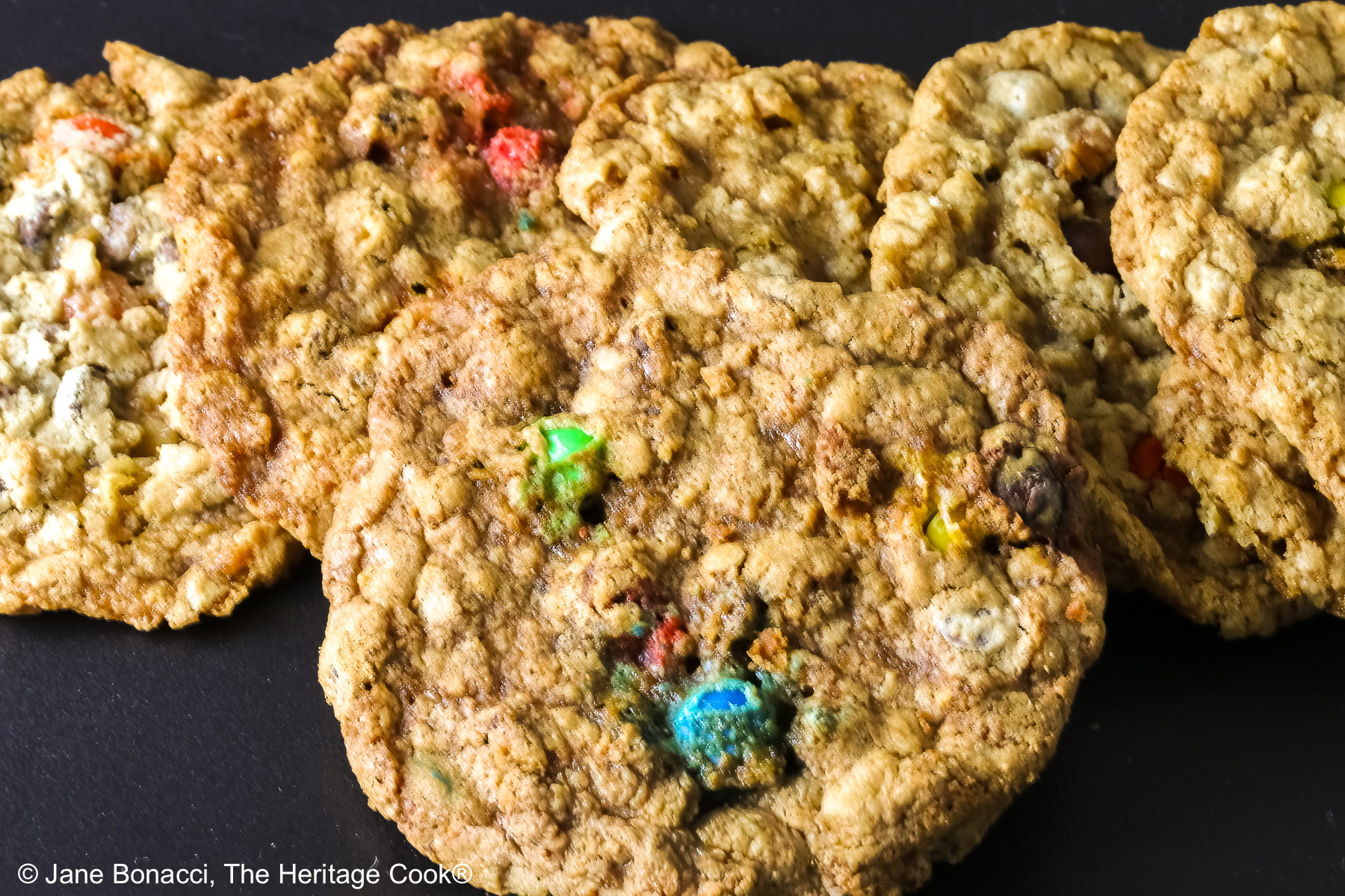 Large oatmeal cookies studded with all kinds of add-ins like coconut, pecans, chocolate chips, peanut butter chips, and M&M candies, laid out on a black rectangular platter with a deep pink cloth in the back and sitting on a mottled gray background © 2026 Jane Bonacci, The Heritage Cook.