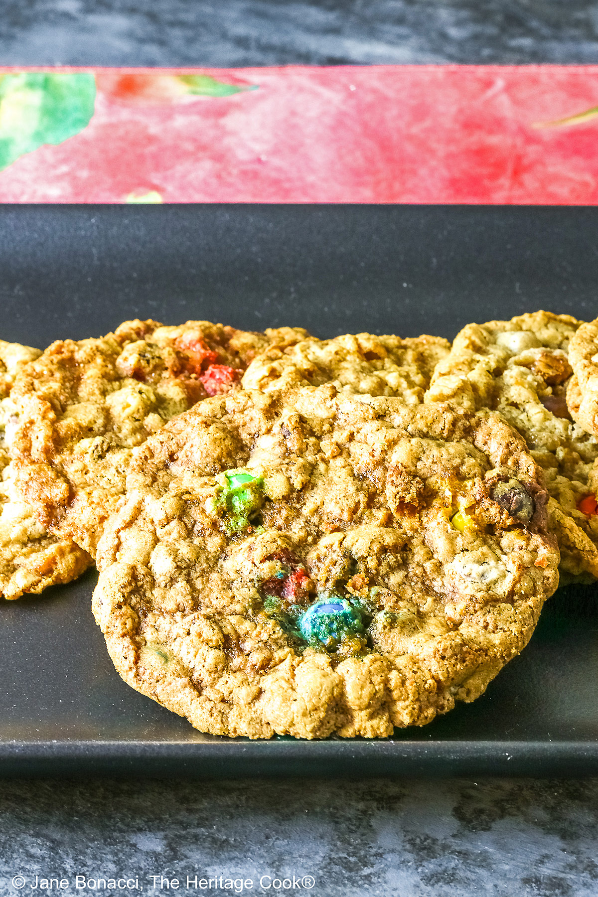 Large oatmeal cookies studded with all kinds of add-ins like coconut, pecans, chocolate chips, peanut butter chips, and M&M candies, laid out on a black rectangular platter with a deep pink cloth in the back and sitting on a mottled gray background © 2026 Jane Bonacci, The Heritage Cook.
