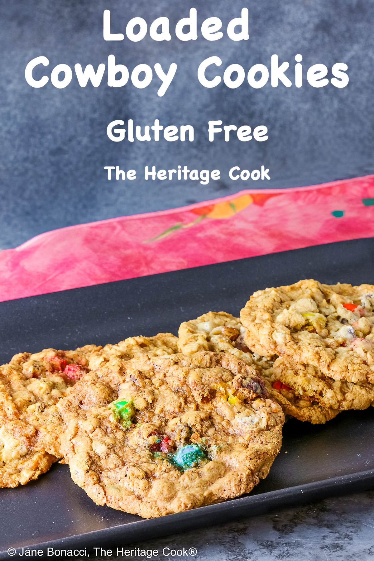 Large oatmeal cookies studded with all kinds of add-ins like coconut, pecans, chocolate chips, peanut butter chips, and M&M candies, laid out on a black rectangular platter with a deep pink cloth in the back and sitting on a mottled gray background © 2026 Jane Bonacci, The Heritage Cook.