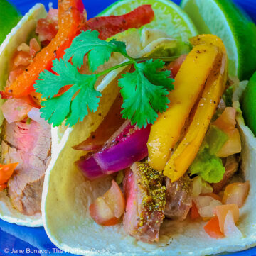 Two One-Pan Steak Fajitas tacos in corn tortillas on a dark blue plate, topped with guacamole, pico de gallo salsa, roasted onions, and bell pepper strips, surrounded by whole limes and lime wedges, with a multi-colored cloth behind and gray background © 2026 Jane Bonacci, The Heritage Cook.