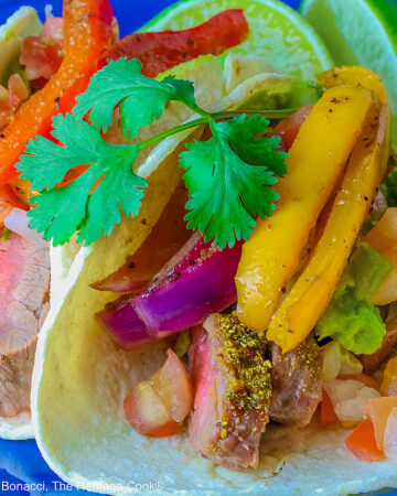 Two One-Pan Steak Fajitas tacos in corn tortillas on a dark blue plate, topped with guacamole, pico de gallo salsa, roasted onions, and bell pepper strips, surrounded by whole limes and lime wedges, with a multi-colored cloth behind and gray background © 2026 Jane Bonacci, The Heritage Cook.
