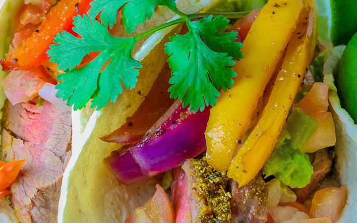 Two One-Pan Steak Fajitas tacos in corn tortillas on a dark blue plate, topped with guacamole, pico de gallo salsa, roasted onions, and bell pepper strips, surrounded by whole limes and lime wedges, with a multi-colored cloth behind and gray background © 2026 Jane Bonacci, The Heritage Cook.