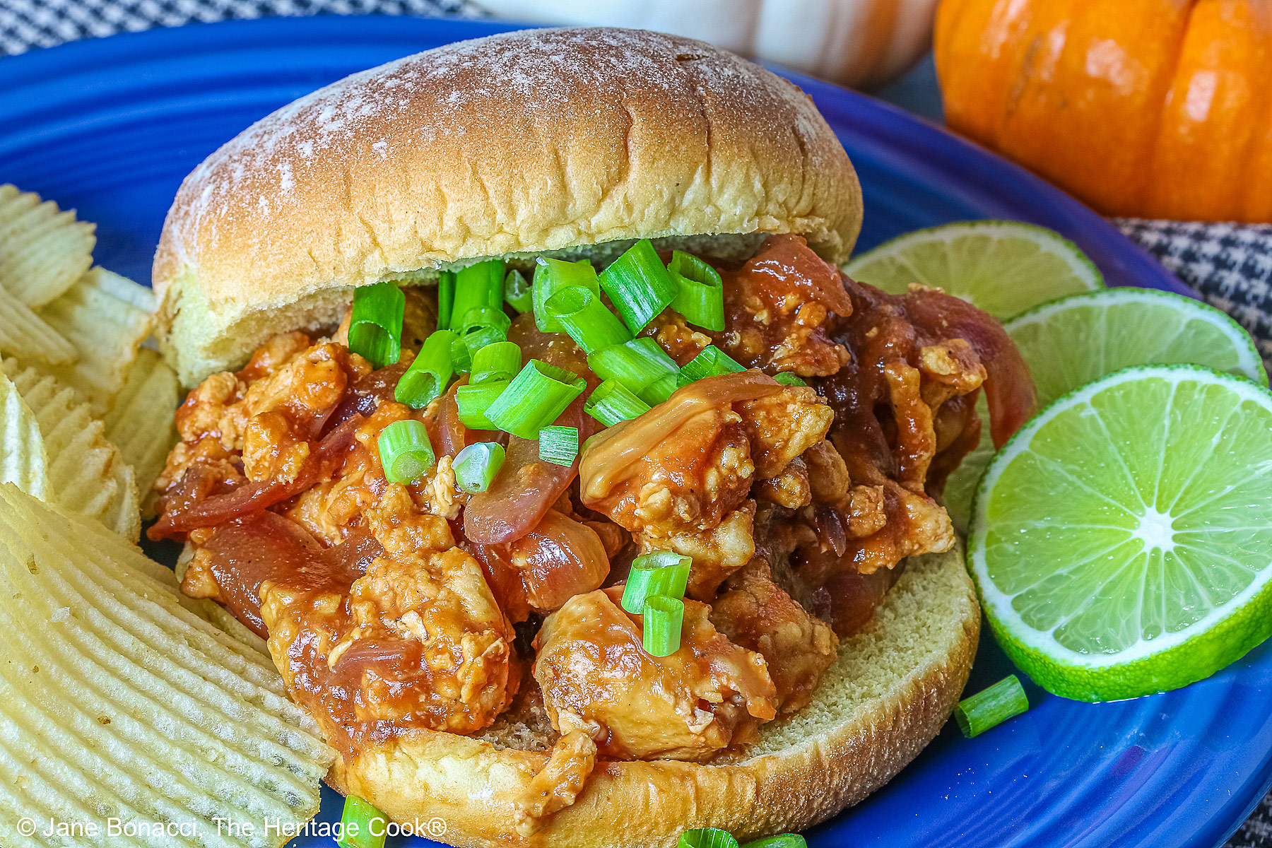 Fully stuffed Asian sloppy joe on a deep blue plate with the top bun tipped on the back, flanked with chips on one side and lime slices on the other and topped with chopped green onions sprinkled over the top and mini pumpkins behind © 2026 Jane Bonacci, The Heritage Cook.