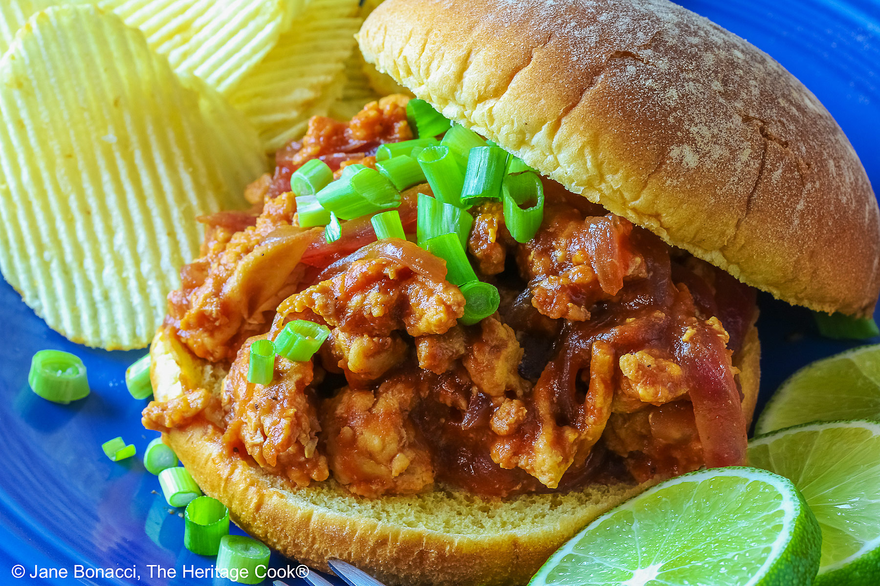 Fully stuffed Asian sloppy joe on a deep blue plate with the top bun tipped on the back, flanked with chips on one side and lime slices on the other and topped with chopped green onions sprinkled over the top and mini pumpkins behind © 2026 Jane Bonacci, The Heritage Cook.