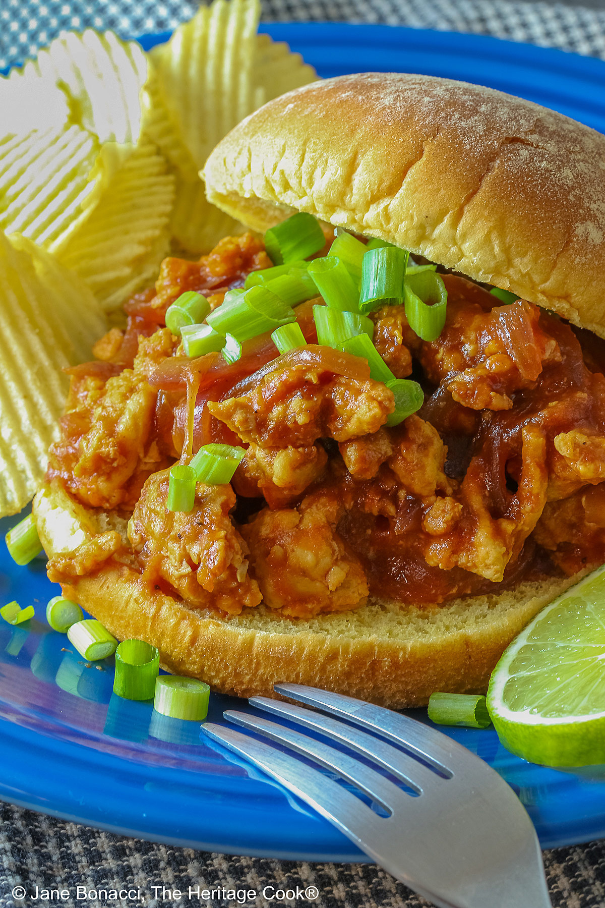 Fully stuffed Asian sloppy joe on a deep blue plate with the top bun tipped on the back, flanked with chips on one side and lime slices on the other and topped with chopped green onions sprinkled over the top and mini pumpkins behind © 2026 Jane Bonacci, The Heritage Cook.