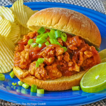 Fully stuffed Asian sloppy joe on a deep blue plate with the top bun tipped on the back, flanked with chips on one side and lime slices on the other and topped with chopped green onions sprinkled over the top and mini pumpkins behind © 2026 Jane Bonacci, The Heritage Cook.