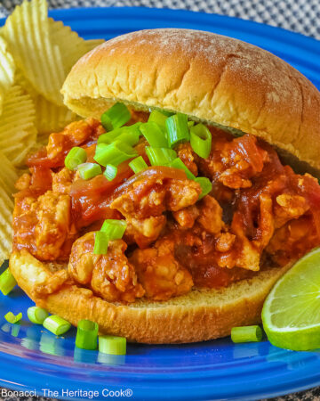 Fully stuffed Asian sloppy joe on a deep blue plate with the top bun tipped on the back, flanked with chips on one side and lime slices on the other and topped with chopped green onions sprinkled over the top and mini pumpkins behind © 2026 Jane Bonacci, The Heritage Cook.