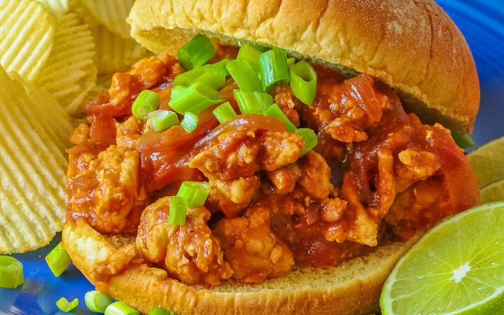 Fully stuffed Asian sloppy joe on a deep blue plate with the top bun tipped on the back, flanked with chips on one side and lime slices on the other and topped with chopped green onions sprinkled over the top and mini pumpkins behind © 2026 Jane Bonacci, The Heritage Cook.