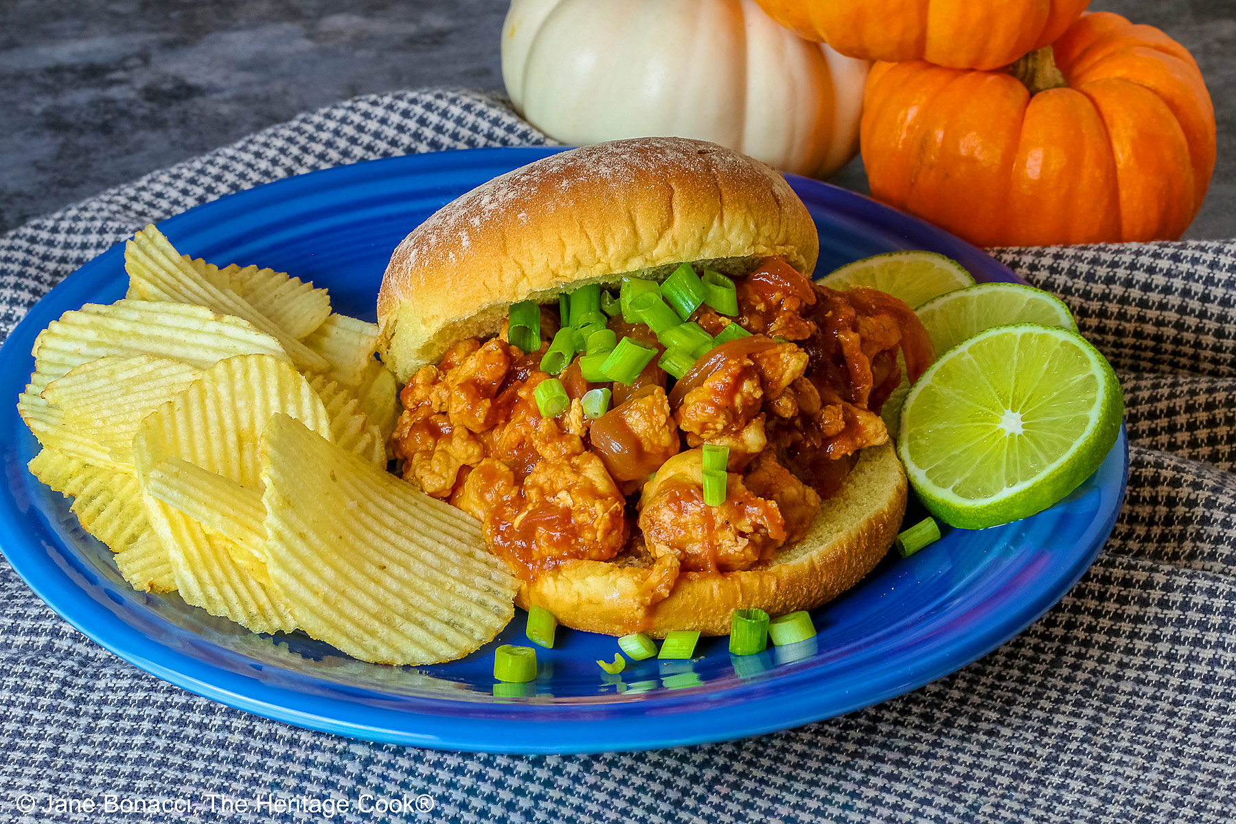 Fully stuffed Asian sloppy joe on a deep blue plate with the top bun tipped on the back, flanked with chips on one side and lime slices on the other and topped with chopped green onions sprinkled over the top and mini pumpkins behind © 2026 Jane Bonacci, The Heritage Cook.