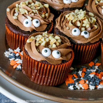 A dark brown plate set on a cream colored plate filled with Nutella Mousse Stuffed Cupcakes each topped with sugar eyes and chopped hazelnuts that look like hair and add a nice texture; there are Halloween themed sugar candies scattered on the plate and a blue cloth gathered behind © 2026 Jane Bonacci, The Heritage Cook.
