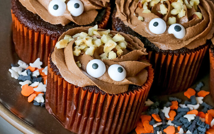 A dark brown plate set on a cream colored plate filled with Nutella Mousse Stuffed Cupcakes each topped with sugar eyes and chopped hazelnuts that look like hair and add a nice texture; there are Halloween themed sugar candies scattered on the plate and a blue cloth gathered behind © 2026 Jane Bonacci, The Heritage Cook.