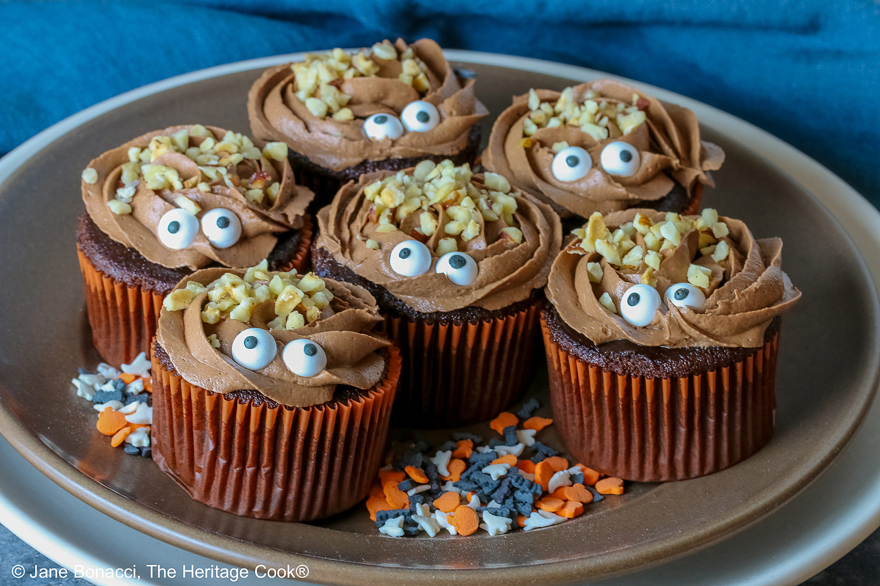 A dark brown plate set on a cream colored plate filled with Nutella Mousse Stuffed Cupcakes each topped with sugar eyes and chopped hazelnuts that look like hair and add a nice texture; there are Halloween themed sugar candies scattered on the plate and a blue cloth gathered behind © 2026 Jane Bonacci, The Heritage Cook.
