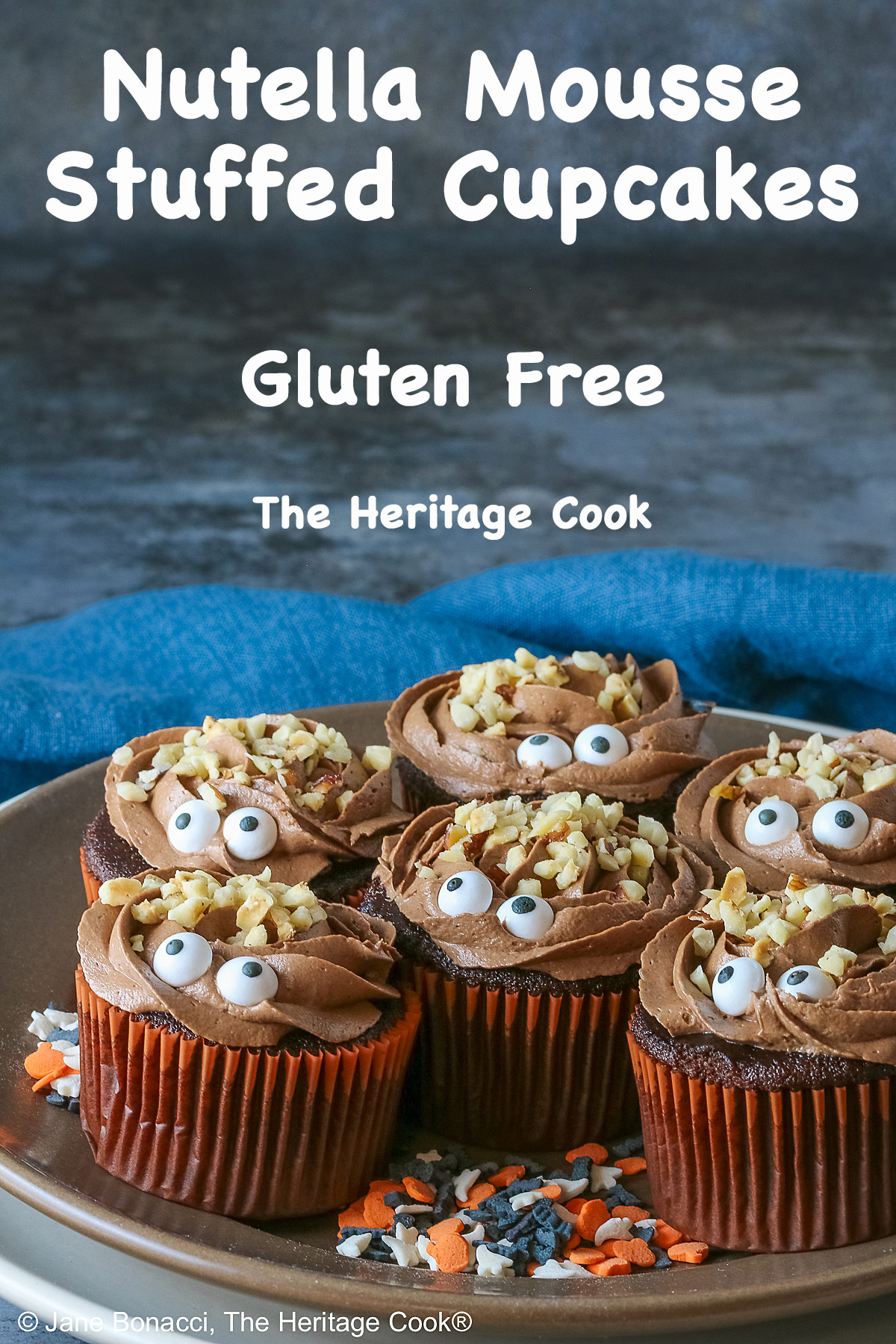 A dark brown plate set on a cream colored plate filled with Nutella Mousse Stuffed Cupcakes each topped with sugar eyes and chopped hazelnuts that look like hair and add a nice texture; there are Halloween themed sugar candies scattered on the plate and a blue cloth gathered behind © 2026 Jane Bonacci, The Heritage Cook.
