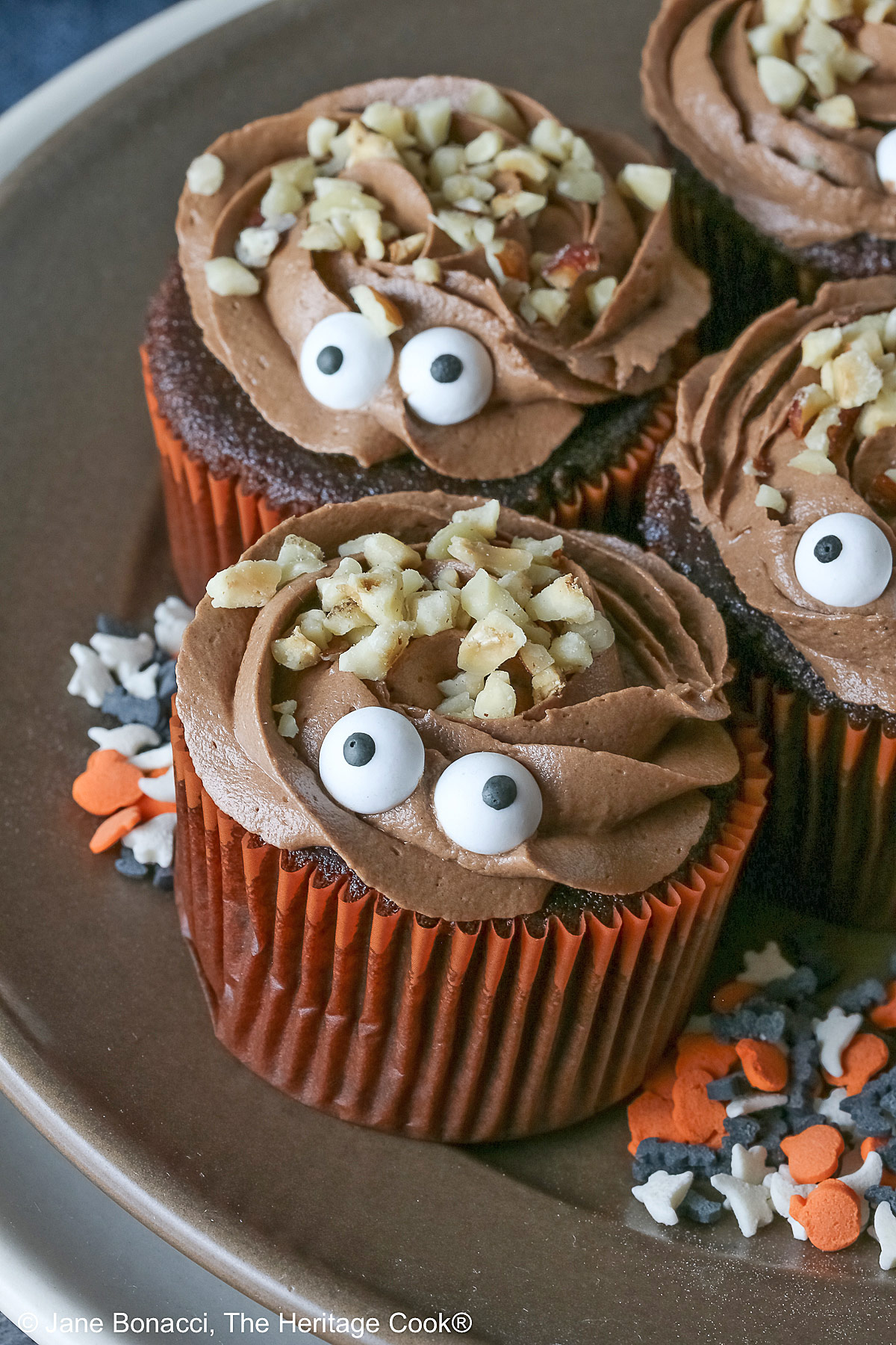 A dark brown plate set on a cream colored plate filled with Nutella Mousse Stuffed Cupcakes each topped with sugar eyes and chopped hazelnuts that look like hair and add a nice texture; there are Halloween themed sugar candies scattered on the plate and a blue cloth gathered behind © 2026 Jane Bonacci, The Heritage Cook.