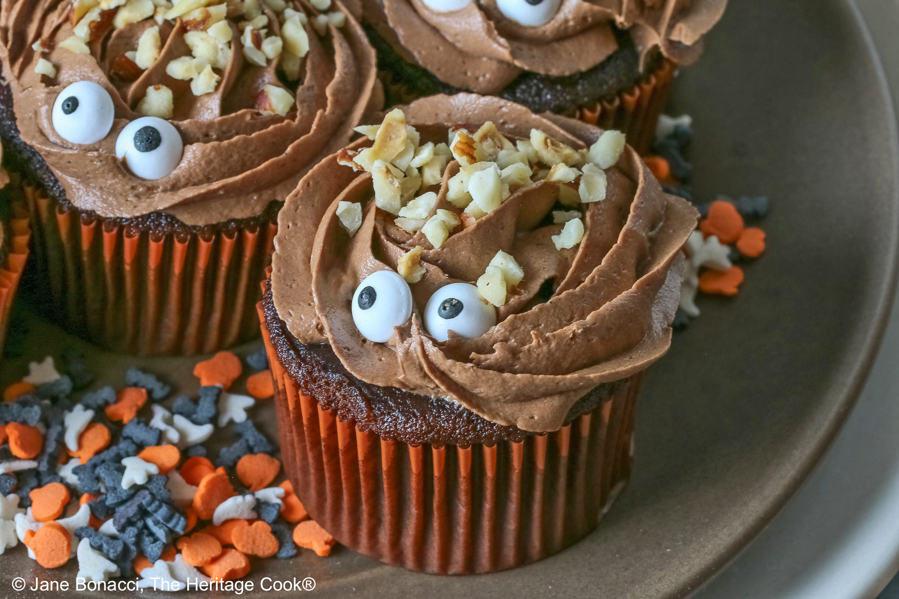 A dark brown plate set on a cream colored plate filled with Nutella Mousse Stuffed Cupcakes each topped with sugar eyes and chopped hazelnuts that look like hair and add a nice texture; there are Halloween themed sugar candies scattered on the plate and a blue cloth gathered behind © 2026 Jane Bonacci, The Heritage Cook.